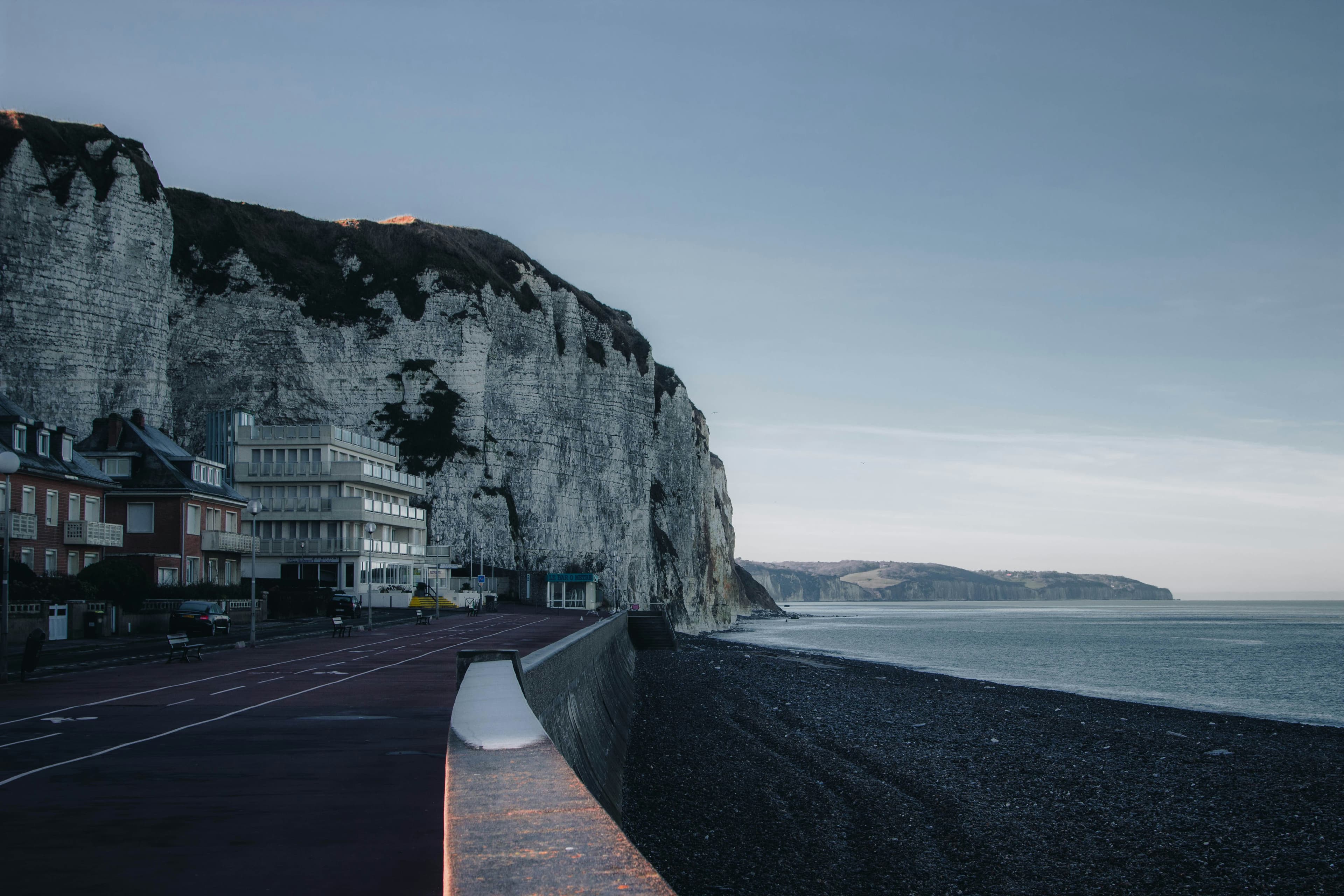 Krijtrotsen bij Dieppe gezien vanaf de kliftop — witte wanden links en rechts, de Kanaalzee in staalgrijs daartussen. Meeuwen als V-vormen in de stijgstroom. Hoge horizon, weinig lucht.