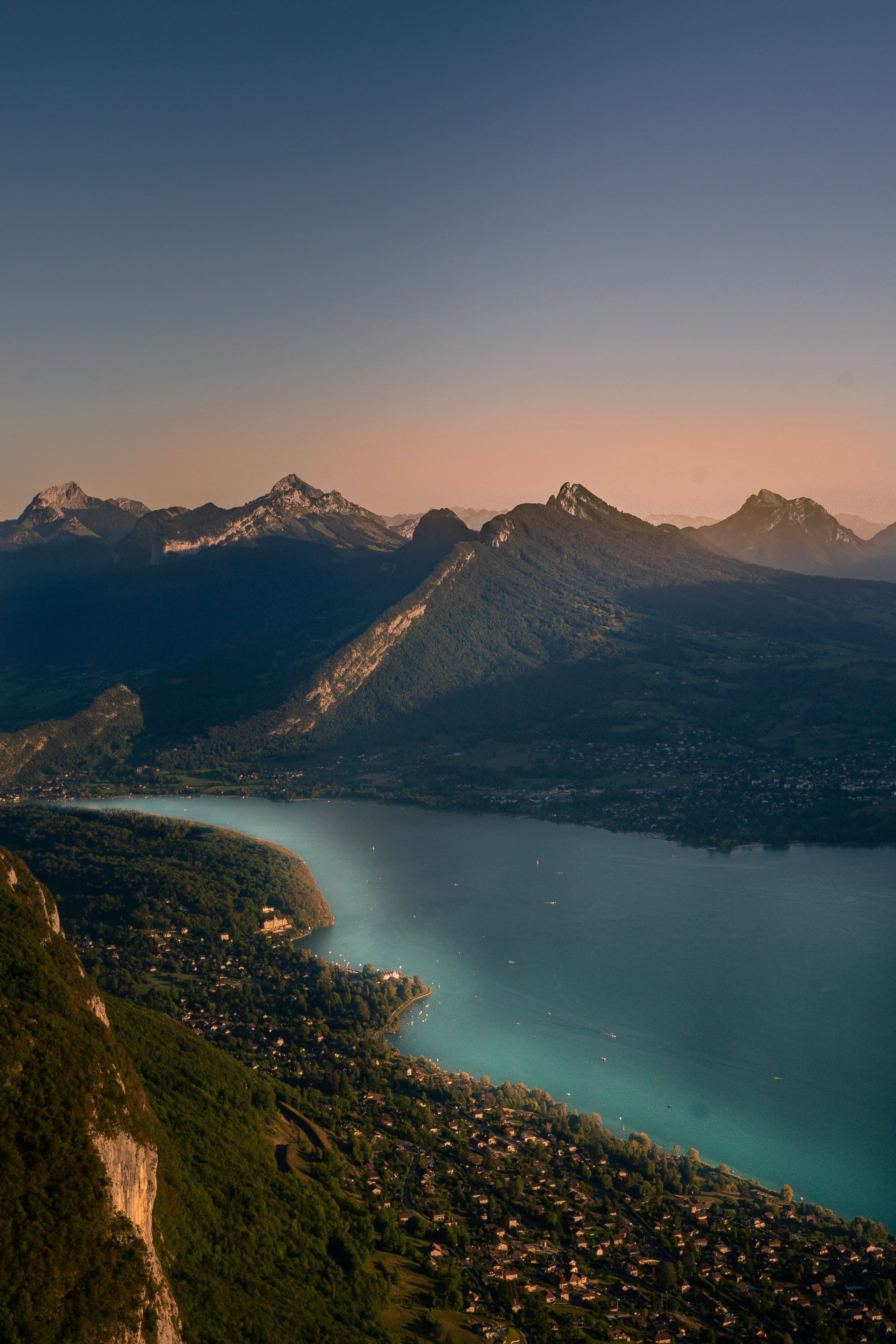 Lac d'Annecy bij vroege ochtend — bergketen weerspiegelt in glas-stil water, ochtendnevel op halve hoogte, bootsteiger als diagonale lijn in de voorgrond. Blauw-grijs toonschaal.